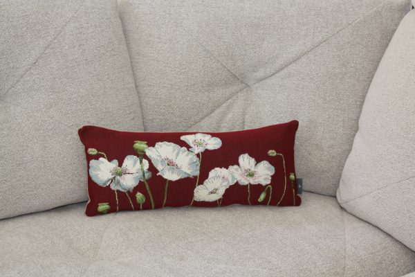Coussin rectangulaire en jacquard rouge orné de coquelicots blancs, sur un canapé gris.