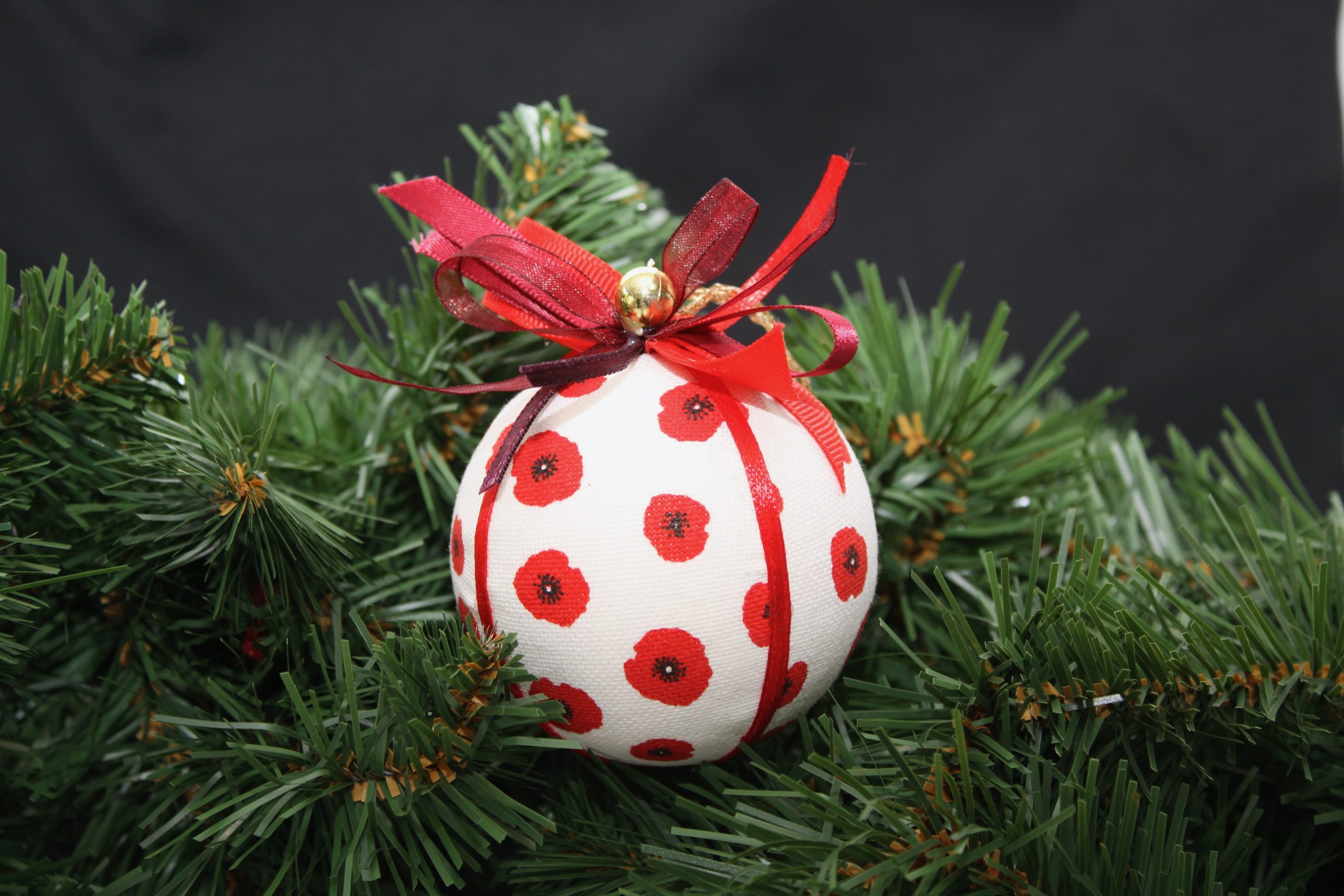 Boule de Noël en tissu blanc ornée de coquelicots rouges, agrémentée de rubans, suspendue parmi des branches de sapin.