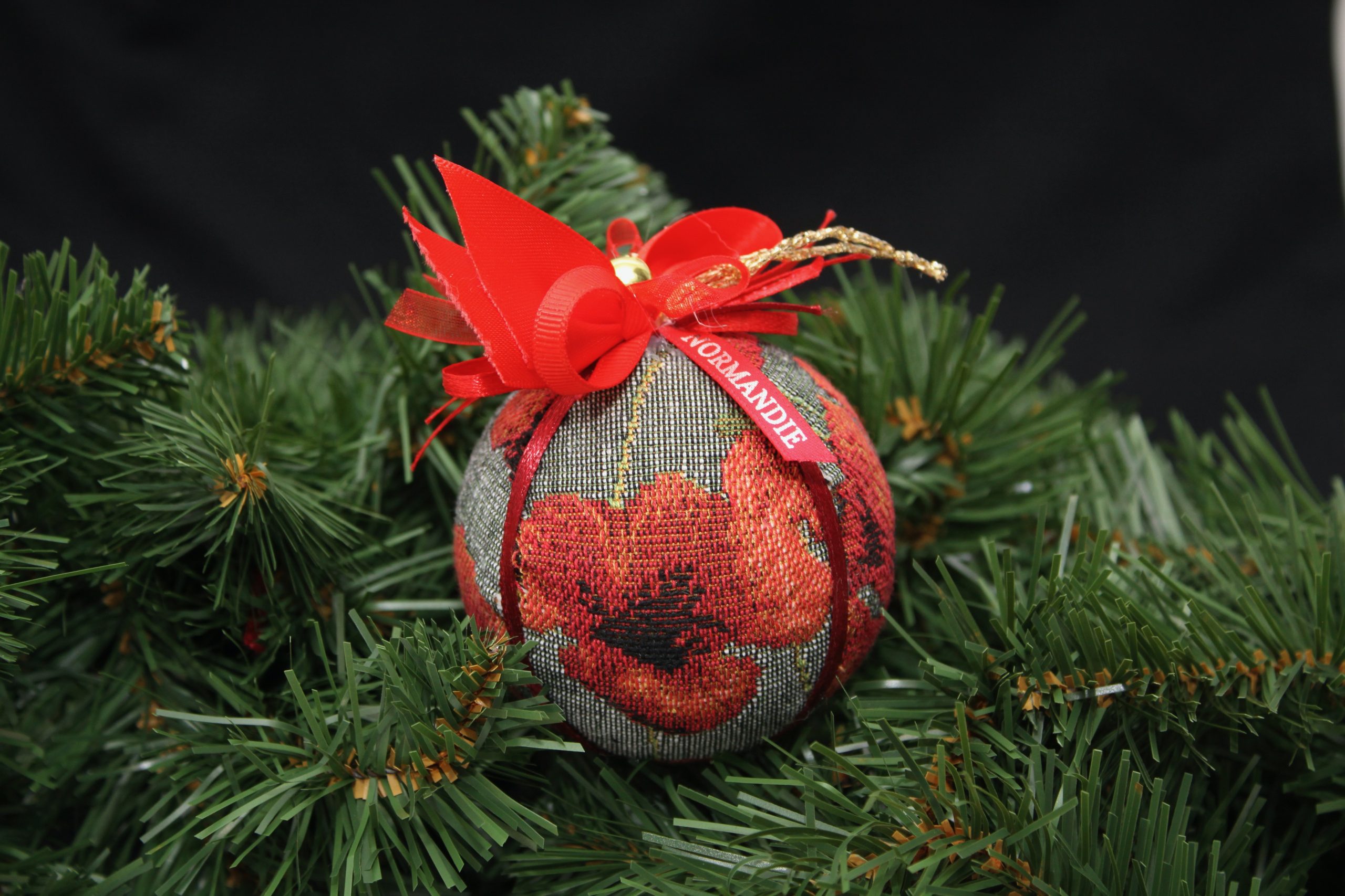 Boule de Noël en tissu jacquard avec motifs de coquelicots, ornée d'un ruban rouge, sur un fond d’aiguilles de sapin.